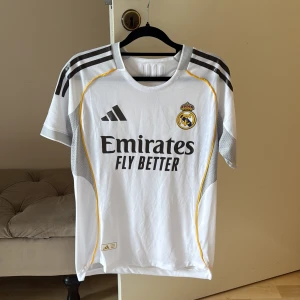 Real Madrid vit matchtröja Adidas S - Säljer en oanvänd Real Madrid fotbollströja från Adidas i storlek S. Tröjan är vit med svarta detaljer, gula linjer och klubbmärke på bröstet. Materialet är lätt och ventilerande polyester, perfekt för träning eller match. Tryck med Emirates Fly Better på framsidan.