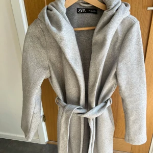 Grå kappa med huva från Zara - Snygg grå kappa från Zara med huva och knytskärp i midjan. Långärmad och i mjukt material som ger en clean och stilren look. Perfekt för dig som gillar minimalistisk stil och vill ha något som funkar till många outfits.