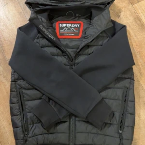 Svart pufferjacka från Superdry - Svart pufferjacka från Superdry Storm Series med huva och dragkedja. Jackan har quiltad kropp, släta svarta ärmar och ribbade muddar. Tillverkad i polyester och bomull, perfekt för kyliga dagar. Snygg logga på bröstet och axeln ger en sportig vibe.