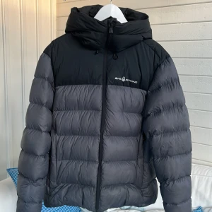 Svart och grå dunjacka Sail Racing - Säljer en dunjacka från Sail Racing i svart och mörkgrått. Jackan har huva, dragkedja framtill och logga på bröstet. Puffig modell som håller dig varm under kalla dagar. Perfekt för dig som gillar stilren och sportig look. Nypris 4200 kr. Liten lagning på höger ärm. 