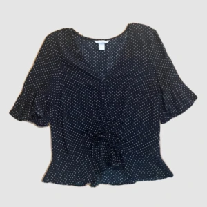 Svart prickig blus med knyt - Supersöt svart blus med vita prickar och v-ringning. Korta, lite fladdriga ärmar och knyt i midjan ger en feminin siluett. Blusen har volangkant nertill och är perfekt för dig som gillar en lekfull men stilren look.