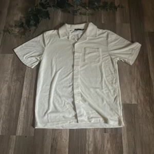 Vit kortärmad skjorta från 157 - Snygg vit skjorta från 157 med korta ärmar och klassisk krage. Skjortan har en bröstficka och knäppning framtill. Tillverkad i mjuk frotté som ger en chill och unik känsla. Perfekt för dig som gillar stilren och enkel design.