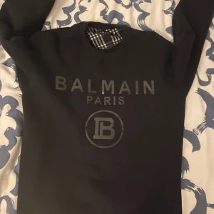 Svart sweatshirt från Balmain Paris - Svart sweatshirt från Balmain Paris med stor logotyp framtill. Tröjan har rund hals och långa ärmar. Insidan har ett rutigt mönster i svart och vitt vid halsen. Perfekt för dig som gillar exklusiva streetwear-vibbar.