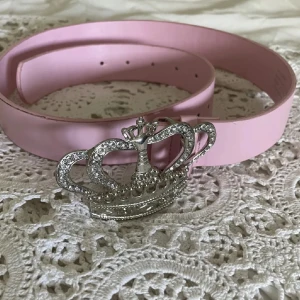 Rosa bälte med krona-spänne - Säljer ett rosa bälte med ett riktigt statement-spänne i form av en glittrande krona med strassdetaljer. Bältet är slätt och har en cool, feminin vibe som passar perfekt till jeans eller kjol. Spännet är i silverfärg och ger en lyxig touch till din outfit. Är lite trasig på vissa delar men syns inte när man har på sig den om du vill se be om bild!!