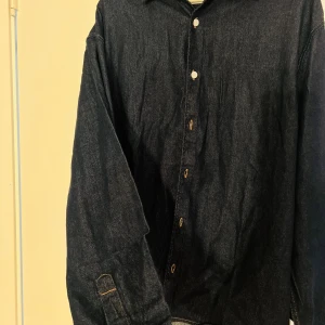 Mörkblå jeansskjorta Jack & Jones - Snygg mörkblå jeansskjorta från Jack & Jones med klassisk krage och kontrastsömmar. Skjortan har långa ärmar och knäppning framtill med vita knappar. Perfekt för dig som gillar en stilren och avslappnad look.