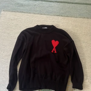 Svart stickad tröja från Ami - Svart stickad tröja från Ami med röd hjärtlogga och bokstaven A på bröstet. Tröjan har rund halsringning, ribbade muddar och lång ärm. Perfekt för dig som gillar stilrena plagg med en twist. Tröjan är oanvänd