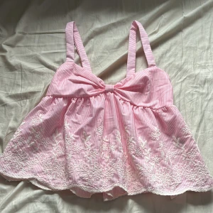 Rosa och vit randig blus - Supersöt blus i rosa och vitt med smala ränder, broderade blommor nertill och en stor rosett framtill. Toppen har breda axelband, smock i ryggen och är tillverkad i lätt polyester. Perfekt för en somrig look! 💕🤍