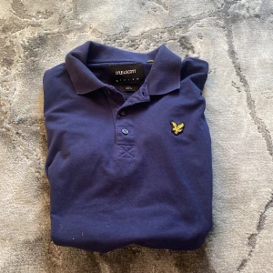 Mörkblå pikétröja från Lyle & Scott - Snygg mörkblå pikétröja från Lyle & Scott med klassisk krage och två knappar framtill. Tröjan har det ikoniska gula fågel-loggan broderad på bröstet. Tillverkad i mjuk bomull och har korta ärmar. Perfekt för en clean och stilren look.