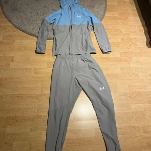 Under Armour träningsset grå/blå - Säljer ett snyggt träningsset från Under Armour med grå byxor och matchande jacka i grått och ljusblått. Jackan har huva och dragkedja framtill, samt logga på bröstet. Perfekt för dig som vill ha en sportig och stilren look.