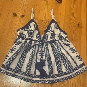 Vit och blå mönstrad linnetopp från SHEIN - Supersöt linnetopp från SHEIN i storlek S med blått och vitt etnoinspirerat mönster. Toppen har smala axelband, v-ringad front och en knytning med tofsar i midjan. Perfekt för dig som gillar boho vibes och vill ha något luftigt till sommaren.