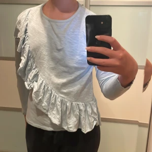 Ljusblå volangtröja från Zara - Supersöt ljusblå långärmad tröja från Zara med volangdetalj som går snett över perfekt för vår och sommar. Enkel att styla till både jeans och kjol för en chill look.
