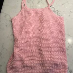 Ljusrosa stickad linnetopp från H&M - Supersöt ljusrosa stickad linnetopp från H&M med smala axelband och virkad känsla. Toppen har en rak passform och diskret ribbad struktur, perfekt för varma dagar. Avslutet nertill har en vågig kant som ger en extra gullig touch.