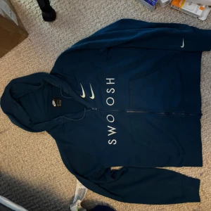 Blå Nike hoodie med Swoosh-tryck - Säljer en marinblå hoodie från Nike med stor Swoosh-logga på ryggen och mindre loggor samt texten 'SWOOSH' på framsidan. Tröjan har huva, dragkedja och två fickor framtill. Perfekt för dig som gillar sportig och stilren streetwear.