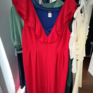 Röd klänning med volang från Redoute - Snygg röd klänning från Redoute med v-ringad hals och bred volang längs axlarna. Klänningen är ärmlös och har ett lätt rynkat tyg som ger en cool struktur. Perfekt för dig som vill sticka ut med färg och volangdetaljer.