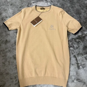 Loro Piana T-shirt  - Snygg beige t-shirt från Loro Piana med broderad logga på bröstet. Rund hals med ribbad kant och mörkblå detaljer. Materialet är mjukt och lyxigt, perfekt för dig som gillar stilrena plagg med exklusiv känsla. Kortärmad och med klassisk passform.
