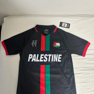 Svart FC Palestine fotbollströja - Snygg svart fotbollströja från FC Palestine med tryck fram och bak. Tröjan har röda och gröna vertikala ränder, röd kant på ärmarna och texten 'PALESTINE' på bröstet samt 'FREE PALESTINE' på ryggen. Tillverkad i lätt och ventilerande material, perfekt för match eller träning. Den är Small men passar även på Medium. Jag själv är Medium och den passade perfekt. 