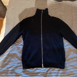 Mörkblå stickad kofta från Zara - Säljer en mörkblå stickad kofta från Zara med hel dragkedja och hög krage. Tröjan är ribbstickad och har långa ärmar, perfekt för lager-på-lager under kyliga dagar. Enkel och stilren design som funkar till det mesta.