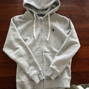 Grå hoodie från Polo Ralph Lauren - Snygg ljusgrå hoodie från Polo Ralph Lauren med dragkedja, huva och vita snören. Klassisk broderad logga på bröstet och mjukt material i bomull och polyester. Perfekt för en chill och stilren look.