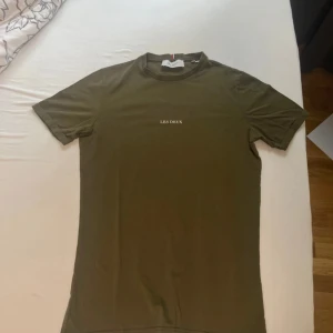 Olivgrön Les Deux t-shirt i bomull - Snygg olivgrön t-shirt från Les Deux med diskret vitt tryck på bröstet. Litet hål som är ganska svår att se men annars helt ny.