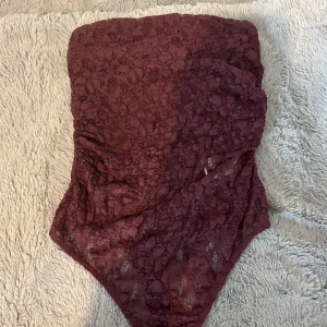 Vinröd spets tubtopp från Bershka - Säljer en vinröd tubtopp från Bershka i spets med blommigt mönster. Toppen är axelbandslös och har bodysuit-modell med tryckknappar nedtill. Det stretchar när man sätter på sig den, kan även visa hur den sitter på!