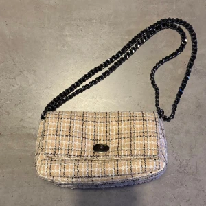 Beck södergard beige rutig axelväska med kedja - Säljer en beige axelväska med rutigt mönster och svart kedjerem. Väskan har en klassisk rektangulär form och stängs med ett snyggt vridlås framtill. Materialet är mjukt och tygigt, vilket ger en trendig och chic känsla.