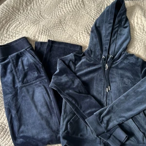 Mörkblå velour hoodie Juicy Couture - Mörkblå hoodie från Juicy Couture i mjuk velour med dragkedja, snörning i huvan och broderad logga på bröstet. Klassisk passform med långa ärmar och ribbade muddar. Perfekt för en chill och trendig look. Pris 1700kr, ordinarie 2000kr. Överdel i storlek xs och byxorna i s!
