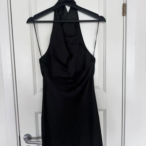 Svart halterneck klänning i satin - Snygg svart kortklänning med halterneck,  draperad halsringning och öppen rygg. Klänningen har en slät, glansig satinlook och tunna axelband. Perfekt för dig som vill ha en stilren och elegant vibe. Enkel att styla med smycken och klackar.