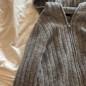 Grå ribbad hoodie från 157, XS - Mysig grå hoodie från 157 i ribbstickat material. Tröjan har huva, dragkedja framtill och långa ärmar. Perfekt för dig som gillar en chill och enkel stil. Passar dig som vill ha en basic men ändå snygg tröja till vardags.