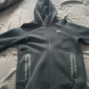 Svart Nike hoodie med dragkedja - Svart hoodie från Nike med hel dragkedja framtill och två fickor med dragkedja. Tröjan har huva med snörning och Nike-logga på bröstet. Materialet är mjukt och känns som en blandning av bomull och polyester, perfekt för chill eller träning.