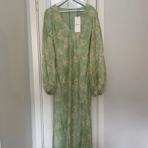 Grön blommig långklänning från Adoore - Pastelgrön långklänning från Adoore med v-ringning och långa puffärmar. Klänningen har ett romantiskt blommigt mönster i vitt och gult, och är tillverkad i ett lätt, glansigt material som ger en lyxig känsla. Perfekt för dig som gillar en feminin och trendig stil. Den är helt oanvänd med alla lappar kvar! 