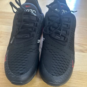 Nike Air Max 270 svarta/röda sneakers - Nike Air Max 270 sneakers i svart med röda detaljer och stor synlig Air-enhet i hälen. Ovandel i mesh för bra andningsförmåga, snörning och röda Swoosh-loggor. Snygg och sportig look med platt sula och bekväm passform.