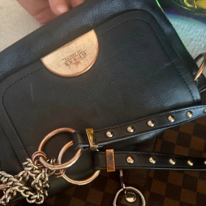 Svart axelväska från Guess - Snygg svart axelväska från Guess i skinn med gulddetaljer och kedja. Väskan har nitar på axelremmen och en stilren design med metallogo framtill. Perfekt för dig som vill ha en trendig och praktisk väska med attityd.