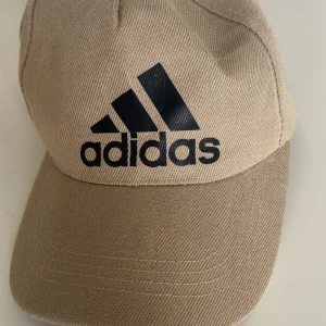 Beige keps från Adidas - Snygg beige keps från Adidas med klassisk svart logga framtill. Kepsen har böjd skärm och justerbar rem baktill för perfekt passform. Tillverkad i tålig bomull som passar året runt. Perfekt accessoar för en sportig och avslappnad stil. Den är köpt i Spanien från en marknad
