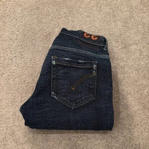 Mörkblå jeans från Dondup - Snygga mörkblå jeans från Dondup, tillverkade i Italien. Klassisk femficksmodell med raka ben och coola slitningar för en avslappnad vibe. Jeansen har kontrastsömmar och en läderpatch med logga bak i midjan. Perfekta för dig som gillar stilrena och trendiga jeans.