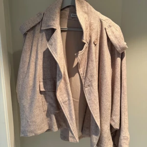 Beige kappa från Alvar of Sweden - Stilren beige kappa från Alvar of Sweden med klassisk krage, axelklaffar och stora fickor framtill. Jackan har en avslappnad passform och är tillverkad i ett mjukt ullblandat tyg som passar perfekt för kyligare dagar.