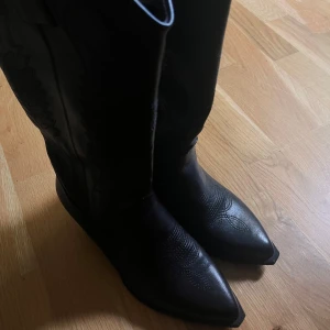 Svarta cowboy boots med spetsig tå - Säljer ett par svarta cowboy boots i skinn med snygga broderade detaljer och spetsig tå. Bootsens höga skaft och klassiska western-silhuett ger en cool vibe. Perfekt för dig som vill sticka ut med en edgy look. Platt sula och låg klack för skön komfort. Storlek 37, men passar de som har 38 också. Använt de en gång och säljer dem, pga att de är för små för mig. :(