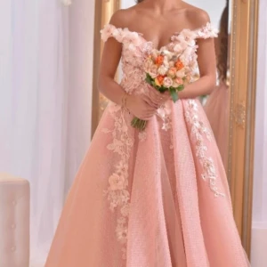 Ljusrosa långklänning med blommor - Drömmig ljusrosa långklänning med offshoulder och 3D-blommor samt spetsdetaljer. Klänningen har en vacker volangkjol och romantiska broderier längs livet. Perfekt för bal eller speciella tillfällen. Köpte för 4000kr