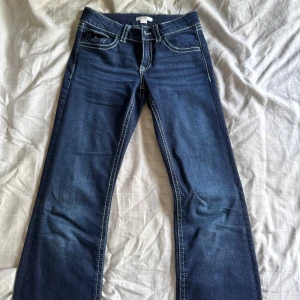 Mörkblå jeans med kontrastsömmar - Snygga mörkblå jeans med tydliga vita kontrastsömmar och klassiska fem fickor. Byxorna har raka ben och coola bakfickor med lock och knapp. Perfekta för dig som gillar en stilren men ändå detaljerad look.