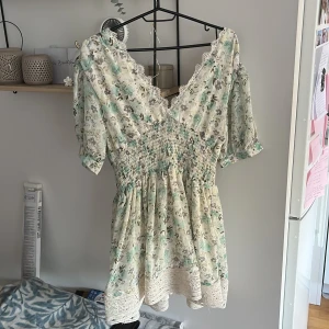 Blommig kortklänning med spetsdetaljer - Supersöt kortklänning i ljusa toner med blommigt mönster i mintgrönt, grått och beige. Klänningen har v-ringad hals med spetskant, puffiga korta ärmar och smock i midjan för en snygg passform. Nederdelen pryds av breda spetsdetaljer. Materialet är 100% polyester.