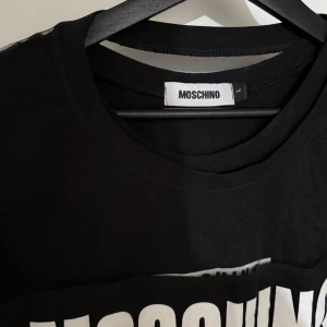 Svart Moschino t-shirt med mesh - Svart t-shirt från Moschino med stor vit logotyp på bröstet. Unik design med transparent mesh på ärmar och sidor, vilket ger en cool och edgy vibe. Rund halsringning och korta ärmar. Perfekt för dig som gillar statement-plagg och streetstyle.