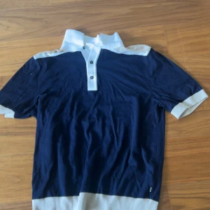 Blå och beige t-shirt från BOSS - Snygg t-shirt från BOSS i storlek S med cool färgblockad design i mörkblått och beige. T-shirten har vit krage, knappslå med tre knappar och korta ärmar med beige muddar. Materialet är mjukt och känns som bomull, perfekt för en chill stil.
