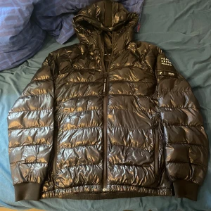 Svart Peak Performance jacka - Säljer en svart, glansig dunjacka från Peak Performance med huva och dragkedja. Jackan har quiltad design, ribbade muddar och två fickor med dragkedja och en innerficka vid vänster bröst. Perfekt för kalla dagar och riktigt snygg streetstil. Den har några defekter, hör av dig om du är intresserad så skickar jag bilder på defekterna. Nypris 2500kr. Pris kan diskuteras😁