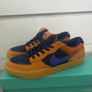Nike SB Force 58 orange/lila sneakers - Nike SB Force 58 sneakers i färgen Monarch/Persian Violet. Skorna har en snygg kombination av orange mocka och mörkblå textil med lila swoosh och detaljer. Perfekta för dig som gillar streetstyle och skate.