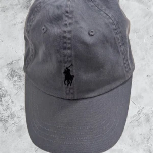 Blå keps från Polo Ralph Lauren - Snygg grå keps från Polo Ralph Lauren med klassisk broderad logga framtill och 'POLO' broderat baktill. Kepsen är i bomull och har justerbar rem med metallspänne där det står 'Polo Ralph Lauren'. Perfekt accessoar för en clean och stilren look.