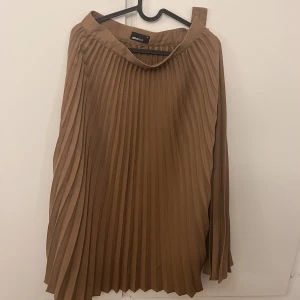 Plisserad brun kjol från Gina Tricot - Säljer en snygg plisserad brun kjol från Gina Tricot i storlek 36. Kjolen har en längre modell och är tillverkad i ett lätt, flowy material som ger ett stilrent intryck. Perfekt att styla med en stickad tröja eller en enkel topp.