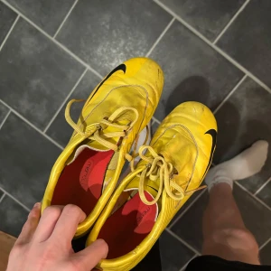  Nike Mercurial Vapor 5 - Snygga gula Nike Mercurial fotbollsskor med svarta detaljer och klassisk swoosh på sidan. Skorna har snörning och dobbar för bra grepp på planen. Insidan är röd och ovandelen är i syntetmaterial för lätt känsla och snabbhet.