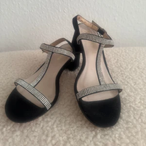 Svarta sandaler med strass och klack - Svarta sandaler med bred klack och remmar täckta av glittrande strass. Skorna har öppen tå och spänne vid ankeln. Utsidan är i mockaliknande material och sulan är ljus. Perfekta för dig som vill sticka ut med lite extra bling. Hade på mig bara en gång inomhus på fest. Lite skavanker, som inte syns när man har skorna på sig, ses sista bild. Innelängd 22,5cm.