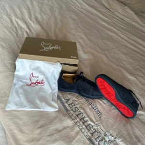 Blåa sneakers från Christian Louboutin - Säljer ett par svarta sneakers från Christian Louboutin med klassisk röd sula och diskret logga. Skorna har snörning och är tillverkade i mocka, vilket ger en lyxig känsla. Perfekta för dig som vill sticka ut med en ikonisk detalj.