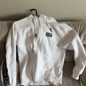 Vit hoodie från Emporio Armani EA7 - Säljer en snygg vit hoodie från Emporio Armani EA7 med dragkedja och huva. Tröjan har svarta logoband längs ärmar och rygg samt en EA7-logga på bröstet. Praktiska fickor framtill och ribbade muddar. Perfekt för en sportig och trendig look.