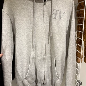 Grå hoodie från Bvaldi med dragkedja - Gråmelerad hoodie från Bvaldi med glittrande logga i strass på bröstet och ryggen. Tröjan har huva med snörning, hel dragkedja och två stora fickor framtill. Perfekt för en chill och avslappnad stil.